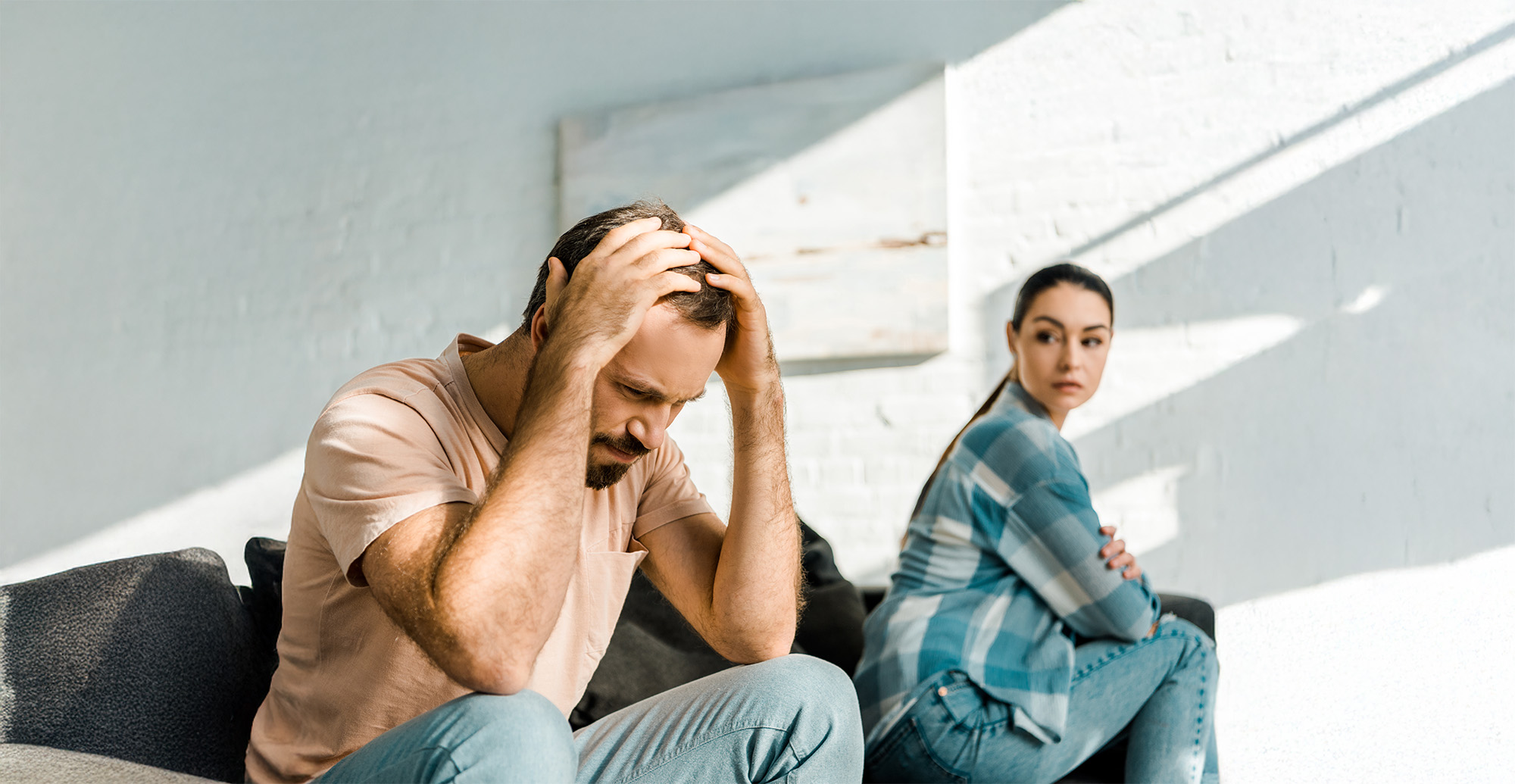 upset man with hands on head sitting on couch after arguing with wife on background