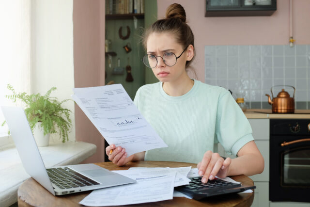 Young frustrated unhappy tired woman with financial troubles, sitting at kitchen table with papers, calculator and laptop computer, reading documents