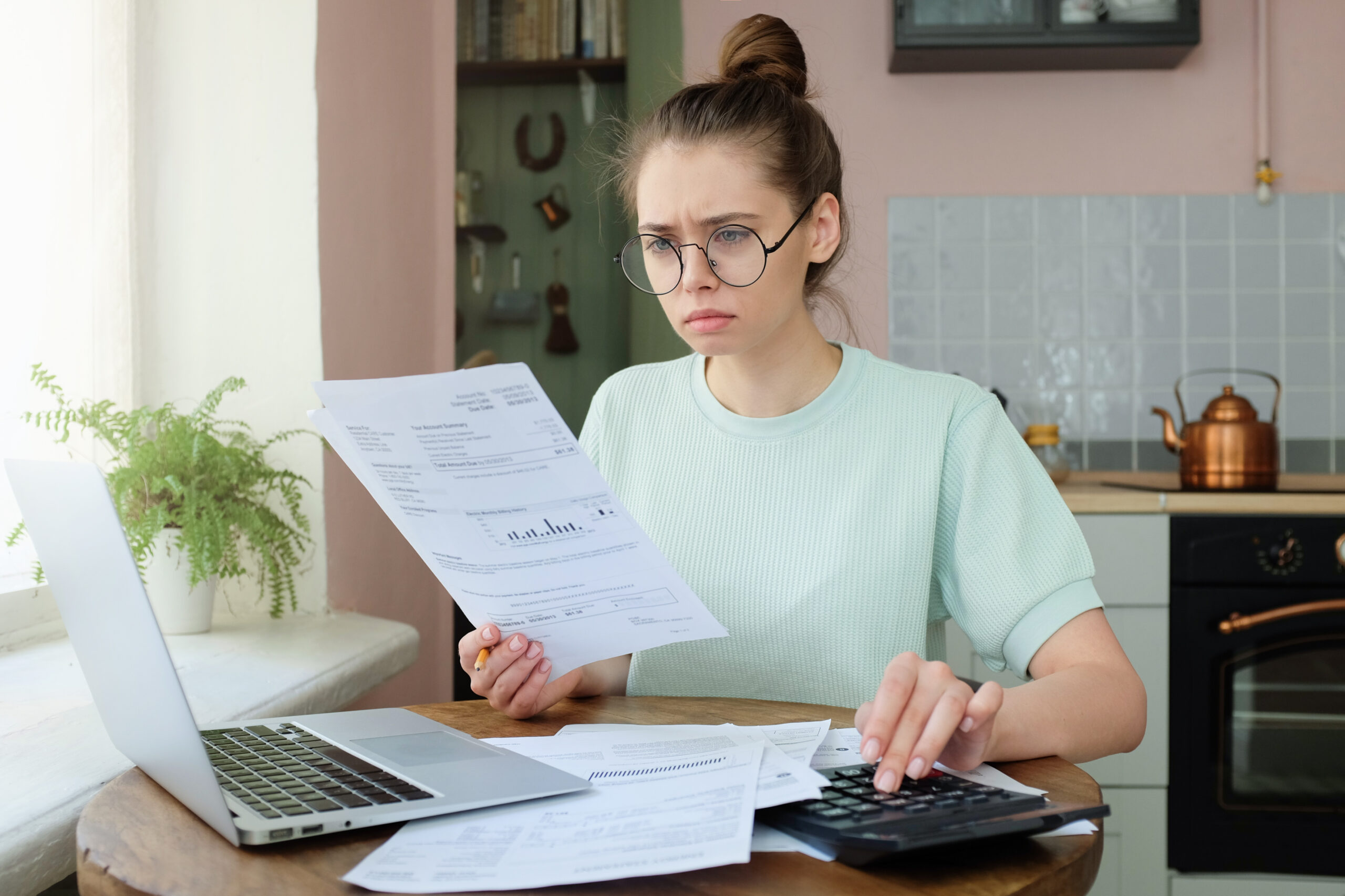 Young frustrated unhappy tired woman with financial troubles, sitting at kitchen table with papers, calculator and laptop computer, reading documents