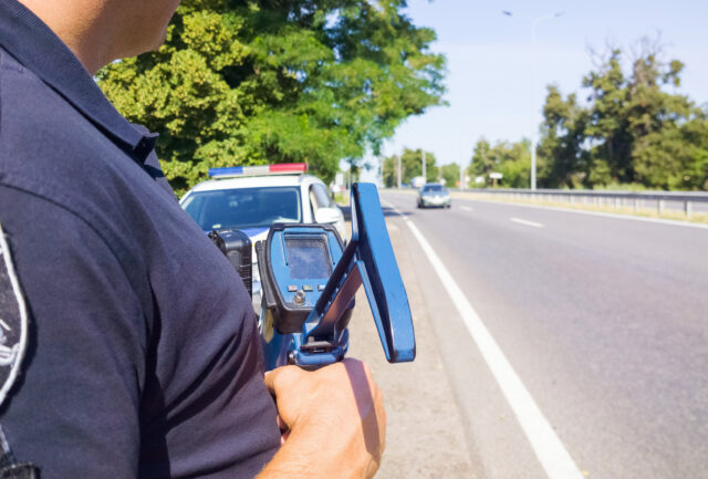 Policeman holding laser speed gun near police car on highway background. Selective focus, part of body.